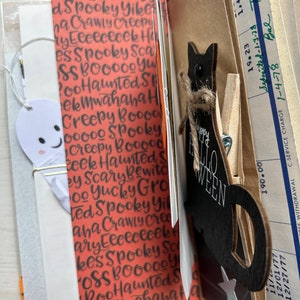 Puede incluir: Una p&aacute;gina de diario con papel naranja y texto negro que incluye Spooky, Haunted y Creepy. Un recorte de fantasma blanco est&aacute; sujeto con un clip dorado. Una silueta de gato de madera negra con un lazo de cuerda y el texto Happy Halloween est&aacute; sujeta a un bolsillo de papel marr&oacute;n que contiene un documento bancario antiguo.