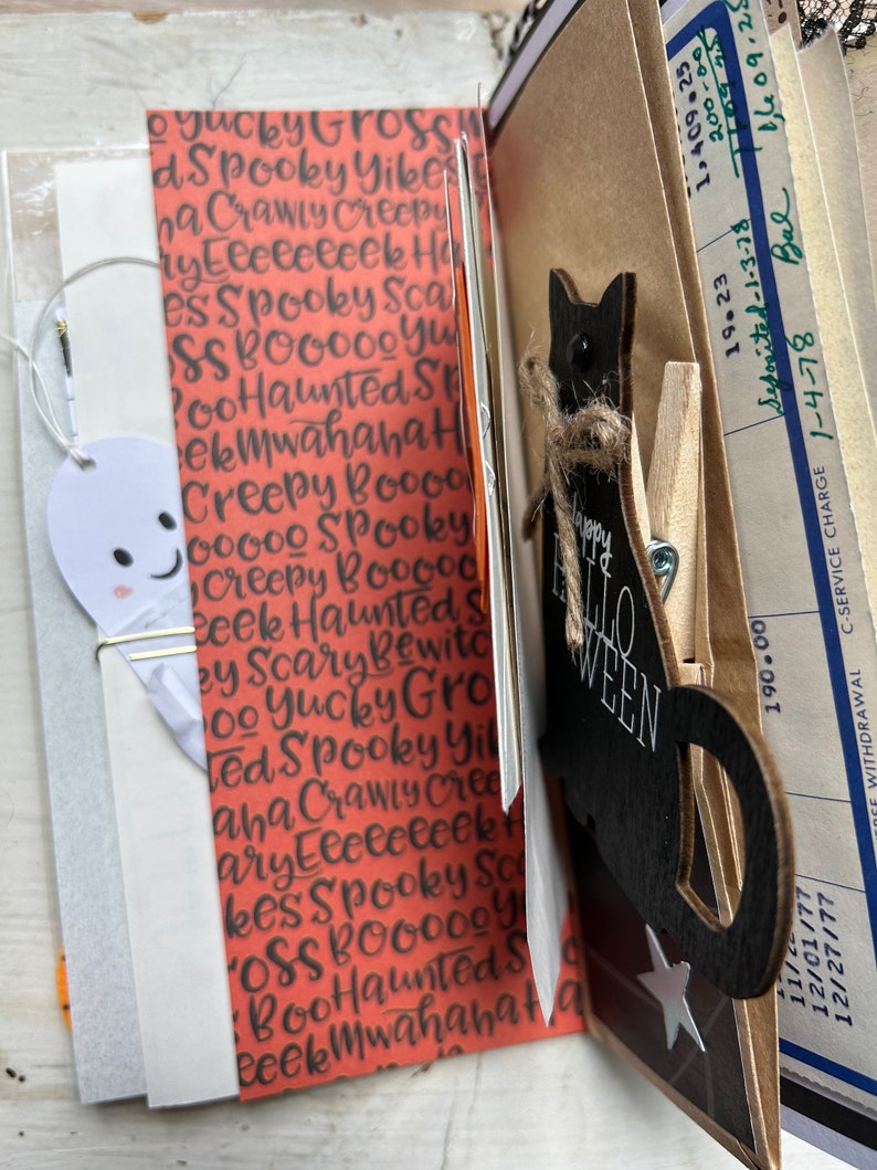Puede incluir: Una p&aacute;gina de diario con papel naranja y texto negro que incluye Spooky, Haunted y Creepy. Un recorte de fantasma blanco est&aacute; sujeto con un clip dorado. Una silueta de gato de madera negra con un lazo de cuerda y el texto Happy Halloween est&aacute; sujeta a un bolsillo de papel marr&oacute;n que contiene un documento bancario antiguo.