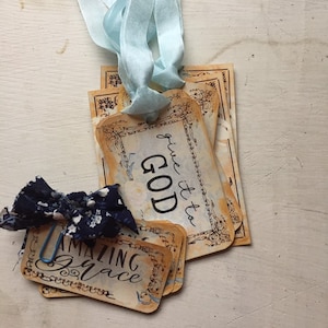 May include: Vintage-style gift tags with the phrases "give it to GOD" and "AMAZING Grace" in elegant script. The tags have a distressed, aged appearance with decorative borders. A blue ribbon and a navy blue bow with a floral pattern add a touch of color.