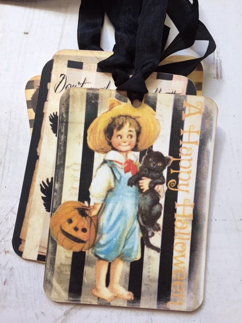 May include: Rectangular gift tags featuring a vintage illustration of a child in a straw hat holding a pumpkin and a black cat. The background has black and white vertical stripes with the text A Happy Halloween printed in orange on the side.