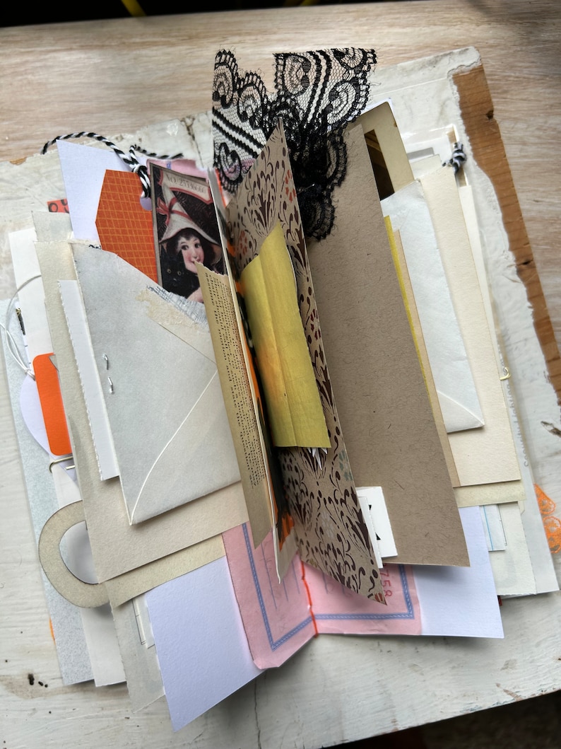 Puede incluir: Un diario artesanal con p&aacute;ginas superpuestas de diversas texturas y colores, incluyendo sobres color crema, cartulina naranja, papel estampado con motivos negros, una ilustraci&oacute;n de estilo vintage y detalles de encaje negro.