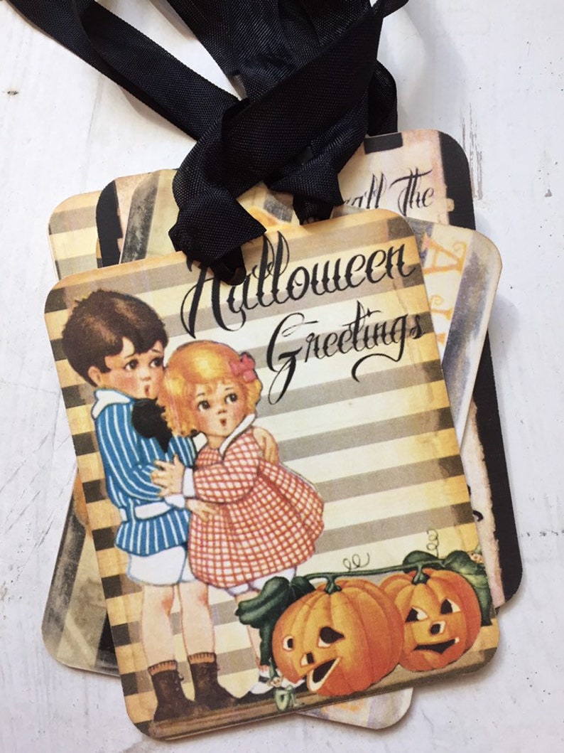 May include: A set of rectangular gift tags with black ribbon ties, featuring a vintage-style illustration of a child in a blue striped shirt and a child in a red checkered dress standing beside two orange jack-o'-lanterns with the text Halloween Greetings.