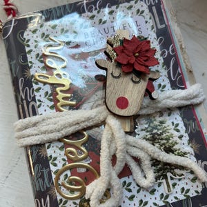 May include: A Christmas-themed journal with a wooden reindeer embellishment. The journal is wrapped with a cream-colored, fluffy cord. Gold lettering spells out "Joy" and "Beautiful Christmas" on the cover. The reindeer has a red nose and a red flower.
