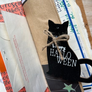 Puede incluir: Una silueta de gato de madera negra con un lazo de cuerda de yute y el texto blanco Happy HALLOWEEN, superpuesta sobre una colecci&oacute;n de ef&iacute;meros de papel vintage, incluyendo un extracto bancario con texto sobre cargos de servicio y un sobre de papel.