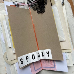 Puede incluir: Un diario artesanal con p&aacute;ginas de papel superpuestas, una encuadernaci&oacute;n central con hilo naranja y un adorno de encaje negro en la parte superior. Una tira de papel blanco con el texto SPOOKY impreso en negro est&aacute; pegada en la parte inferior.
