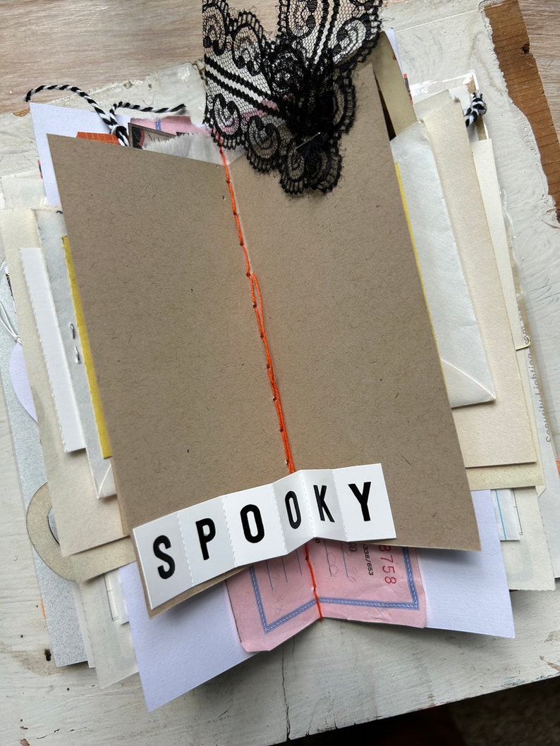 Puede incluir: Un diario artesanal con p&aacute;ginas de papel superpuestas, una encuadernaci&oacute;n central con hilo naranja y un adorno de encaje negro en la parte superior. Una tira de papel blanco con el texto SPOOKY impreso en negro est&aacute; pegada en la parte inferior.