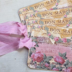 May include: A stack of vintage-style gift tags with a pink ribbon. The tags feature the text "BON MARCHL" and "PARIS" with floral and architectural designs in shades of pink, beige, and green.