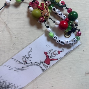 May include: A white rectangular bookmark featuring a black and white illustration of the Grinch and his dog Max. The bookmark is attached to a black cord adorned with various colorful beads, including red, green, and white spheres, and lettered beads spelling GRINCH.