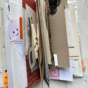 Puede incluir: Un diario artesanal con p&aacute;ginas superpuestas de papel estampado, cartulina y ef&iacute;mera. El libro incluye un recorte con forma de fantasma, una pinza de madera, encaje negro y papel naranja con el texto impreso Spooky, Creepy, Boo.