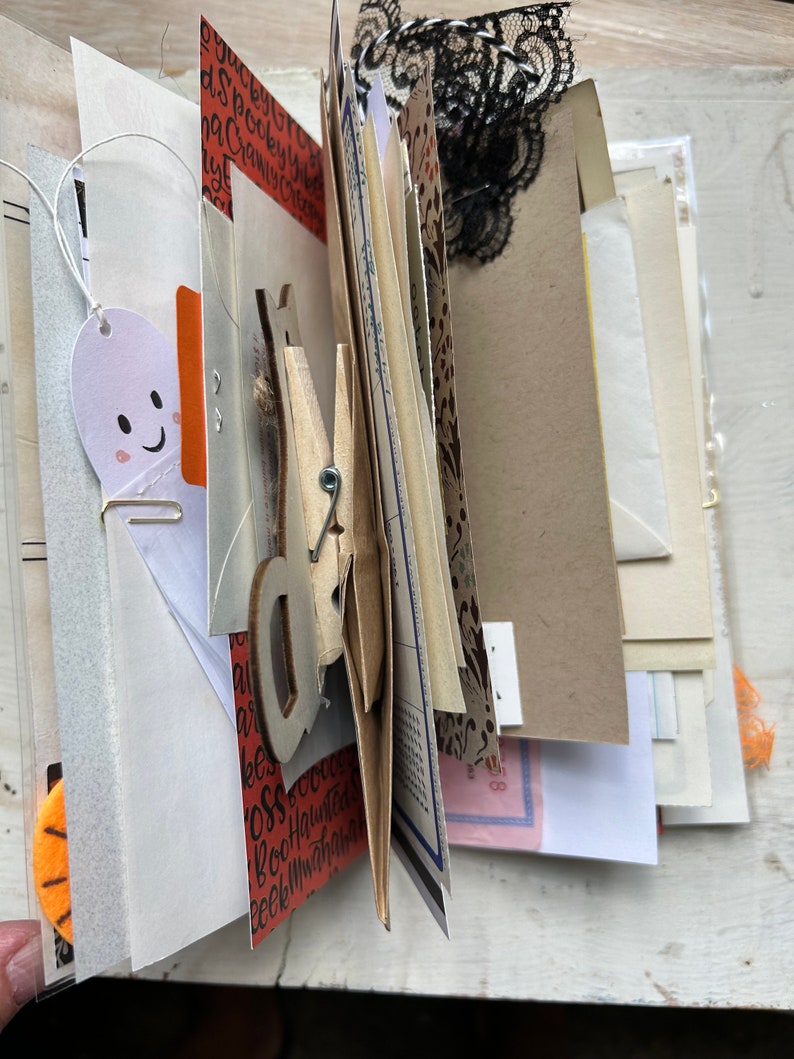 Puede incluir: Un diario artesanal con p&aacute;ginas superpuestas de papel estampado, cartulina y ef&iacute;mera. El libro incluye un recorte con forma de fantasma, una pinza de madera, encaje negro y papel naranja con el texto impreso Spooky, Creepy, Boo.