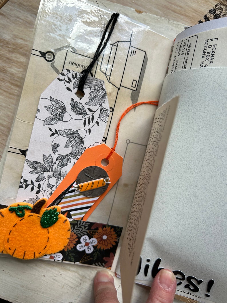 Puede incluir: Una p&aacute;gina de &aacute;lbum de recortes con una etiqueta floral en blanco y negro con cuerda negra, una etiqueta naranja con un adorno de caramelo, un recorte de calabaza de fieltro y una tira de papel floral. Incluye fragmentos de texto y la palabra Vibes!.