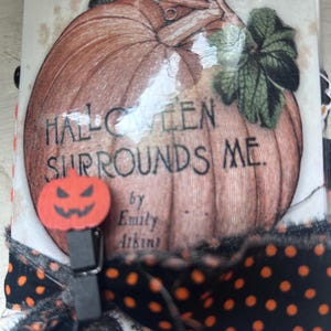 Puede incluir: Una cubierta de libro rectangular con una ilustraci&oacute;n de una gran calabaza naranja y el texto HALLOWEEN SURROUNDS ME de Emily Atkins. Un peque&ntilde;o clip de madera naranja con forma de calabaza est&aacute; sujeto al lateral, decorado con una cinta negra de lunares naranjas.