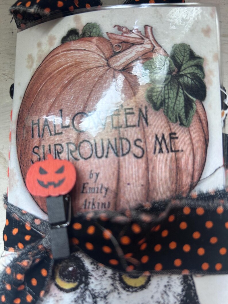 Puede incluir: Una cubierta de libro rectangular con una ilustraci&oacute;n de una gran calabaza naranja y el texto HALLOWEEN SURROUNDS ME de Emily Atkins. Un peque&ntilde;o clip de madera naranja con forma de calabaza est&aacute; sujeto al lateral, decorado con una cinta negra de lunares naranjas.