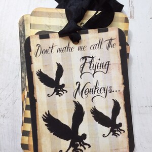 May include: A rectangular paper tag with a black ribbon, featuring a beige and black striped background. The text reads "Don't make me call The Flying Monkeys..." in a black script font, accompanied by three black silhouettes of winged monkeys.