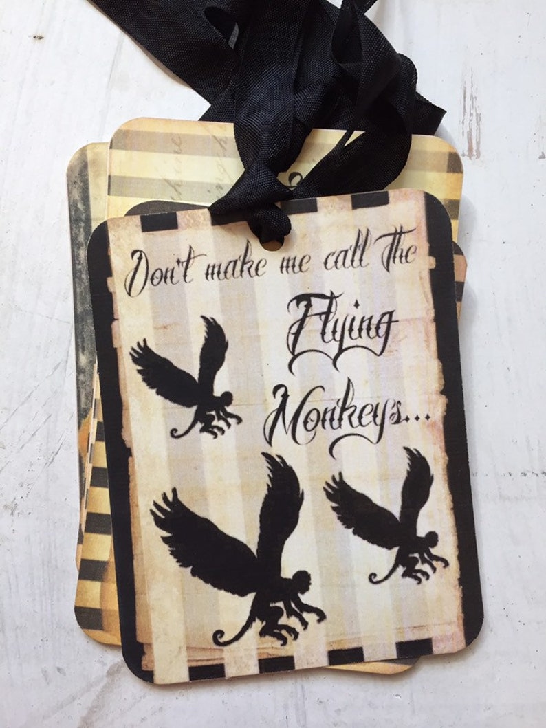 May include: A rectangular paper tag with a black ribbon, featuring a beige and black striped background. The text reads "Don't make me call The Flying Monkeys..." in a black script font, accompanied by three black silhouettes of winged monkeys.