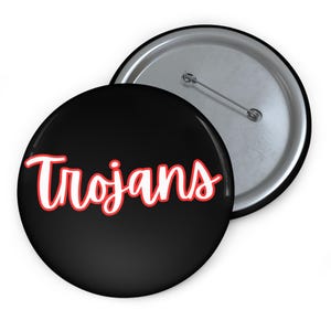 May include: Two round black buttons with a silver backing. One button displays the word "Trojans" in white script with a red outline. The other button is visible in the background.