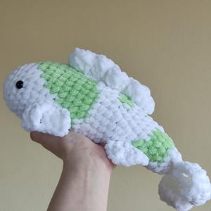 May include: A handmade, plush koi fish in white and light green. The soft, crocheted fish has a rounded body, a small black eye, and fluffy fins and tail. The koi is approximately 12 inches long.