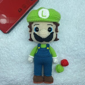 May include: A handmade crochet doll of the character Luigi from the Super Mario Bros. series. The doll features a green hat with a white "L", a tan face, a black mustache, and blue overalls. The doll is approximately 8 inches tall.