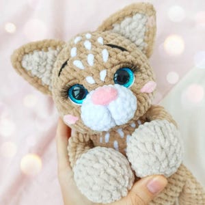 May include: A handmade crochet cat doll with a tan body, white spots, and large blue eyes. The cat has a pink nose and cheeks, with white and tan ears. The doll is soft and plush, with a detailed design.