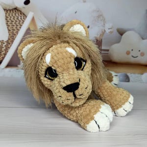 Puede incluir: Un juguete de peluche con forma de le&oacute;n en tonos beige y marr&oacute;n, con una melena esponjosa y detalles blancos en las patas y orejas. El le&oacute;n tiene ojos grandes y expresivos con detalles dorados y una nariz negra. El juguete est&aacute; tumbado sobre una superficie clara.