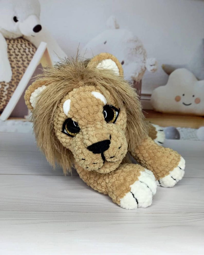 Puede incluir: Un juguete de peluche con forma de le&oacute;n en tonos beige y marr&oacute;n, con una melena esponjosa y detalles blancos en las patas y orejas. El le&oacute;n tiene ojos grandes y expresivos con detalles dorados y una nariz negra. El juguete est&aacute; tumbado sobre una superficie clara.