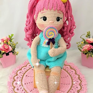 Puede incluir: Muñeca de ganchillo hecha a mano con cabello rizado rosa, vestida con un pelele turquesa y sosteniendo una piruleta colorida. La muñeca tiene un tono de piel claro y está sentada sobre una alfombra de ganchillo rosa.