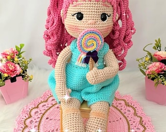 May include: A handmade crochet doll with pink curly hair, wearing a turquoise romper and holding a colorful lollipop. The doll has a light skin tone and is sitting on a pink crocheted mat.