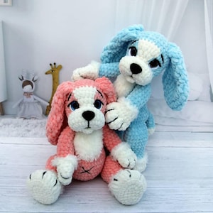 May include: Two plush dog toys. One is pink and white, the other is blue and white. Both have black eyes and noses. The pink dog is sitting, while the blue dog is standing behind it. The toys are made of a soft, fuzzy material.
