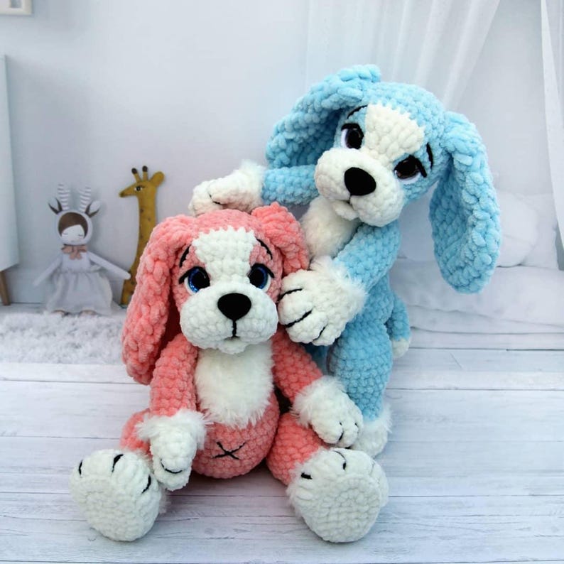 May include: Two plush dog toys. One is pink and white, the other is blue and white. Both have black eyes and noses. The pink dog is sitting, while the blue dog is standing behind it. The toys are made of a soft, fuzzy material.