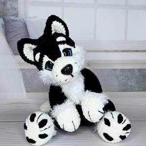 K&ouml;nnte beinhalten: Ein handgefertigtes Husky-Pl&uuml;schtier mit schwarz-wei&szlig;em Fell, blauen Augen und einer flauschigen wei&szlig;en Brust. Der Hund sitzt und zeigt seine schwarzen Pfoten. Das Spielzeug ist aus weichem, strukturiertem Garn gefertigt.