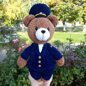 Puede incluir: Un oso de peluche marrón hecho a mano que lleva un uniforme azul marino con botones dorados y un sombrero a juego. El oso tiene un hocico blanco y está hecho de hilo suave y texturizado. Un artículo artesanal y lindo.