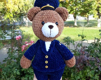 May include: A handmade brown teddy bear wearing a navy blue uniform with gold buttons and a matching hat. The bear has a white muzzle and is made of soft, textured yarn. The bear is a cute, handcrafted item.