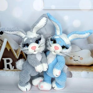May include: Two handmade crochet bunny rabbits. One is gray with white accents, the other is light blue with white accents. Both have long eyelashes, pink noses, and white paws. They are standing close together.