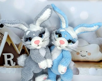 May include: Two handmade crochet bunny rabbits. One is gray with white accents, the other is light blue with white accents. Both have long eyelashes, pink noses, and white paws. They are standing close together.