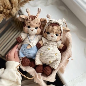 Puede incluir: Dos muñecos de jirafa hechos a mano con ganchillo. Uno lleva un conjunto azul claro, el otro un conjunto color crema con detalles florales. Ambos muñecos tienen detalles marrones y beige, incluyendo sus pezuñas y manchas. Están metidos en una cesta tejida.