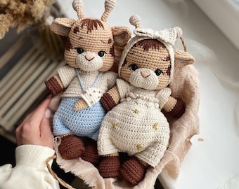 May include: Two handmade crochet giraffe dolls. One doll wears a light blue outfit, the other a cream-colored outfit with floral accents. Both dolls have brown and tan details, including their hooves and spots. They are nestled in a woven basket.