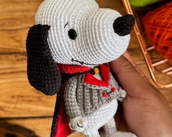 May include: Crocheted Snoopy figure in a vampire costume. The handmade plushie features a white body, black ears and nose, and a gray and red outfit. The figure is approximately 6 inches tall.