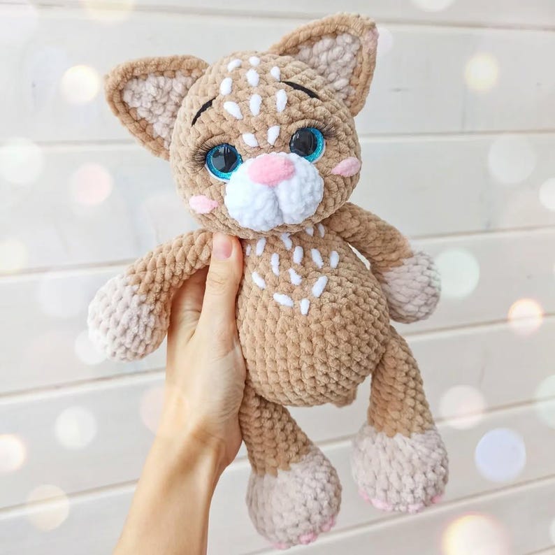 May include: A handmade crochet cat plushie in shades of tan and cream. The cat has blue eyes, a pink nose, and white accents. The plushie is soft and cuddly, perfect for a child's toy or nursery decor.