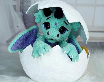 May include: A turquoise crocheted dragon emerging from a white eggshell. The dragon has purple wings, black horns, and large blue eyes. A small gem adorns its forehead. The eggshell is textured and sits on a white surface.