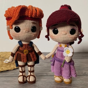 Puede incluir: Dos muñecos de ganchillo hechos a mano, uno con pelo naranja y un atuendo marrón, el otro con pelo burdeos y un vestido morado. Se dan la mano. Un muñeco sostiene una flor blanca.