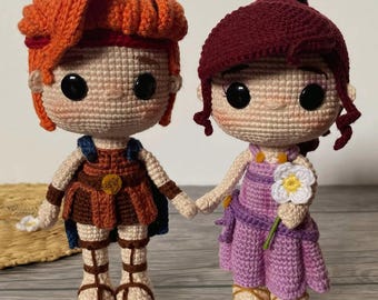 May include: Two crocheted dolls, one with orange hair and a brown outfit, the other with burgundy hair and a purple dress, holding hands. The dolls have large black eyes and are handmade. One doll holds a white flower.