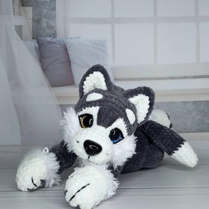 K&ouml;nnte beinhalten: Ein handgefertigtes Husky-Pl&uuml;schtier in Grau, Wei&szlig; und Schwarz. Das Stofftier hat ein goldenes und ein blaues Auge, eine schwarze Nase und wei&szlig;e Pfoten. Der Husky liegt auf einer hellen Oberfl&auml;che.