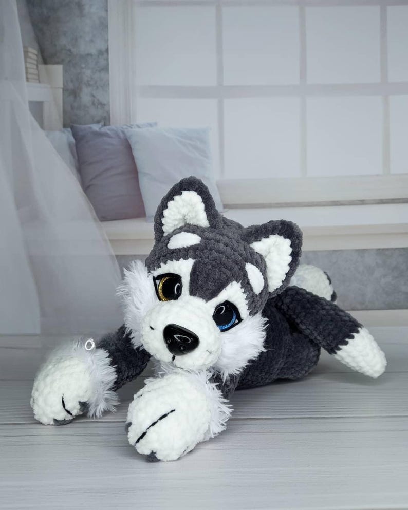 K&ouml;nnte beinhalten: Ein handgefertigtes Husky-Pl&uuml;schtier in Grau, Wei&szlig; und Schwarz. Das Stofftier hat ein goldenes und ein blaues Auge, eine schwarze Nase und wei&szlig;e Pfoten. Der Husky liegt auf einer hellen Oberfl&auml;che.