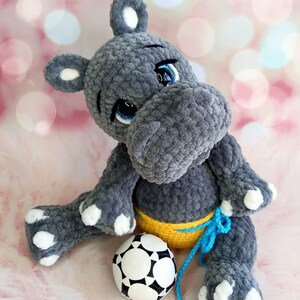 May include: A gray crocheted hippo plush toy with white accents on its ears, feet, and hands. The hippo has blue eyes and is wearing a yellow and blue outfit. A black and white soccer ball is next to the hippo.