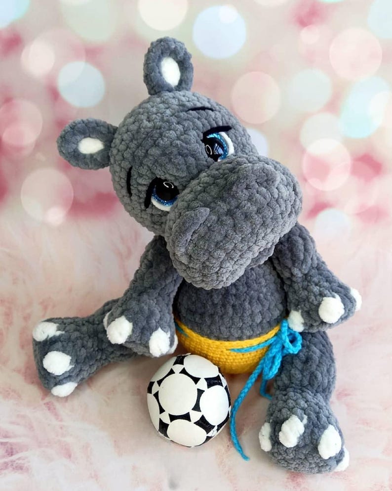 May include: A gray crocheted hippo plush toy with white accents on its ears, feet, and hands. The hippo has blue eyes and is wearing a yellow and blue outfit. A black and white soccer ball is next to the hippo.