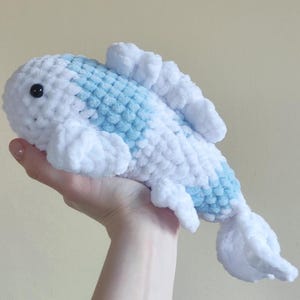 May include: A handmade, plush fish toy in white and light blue. The soft, crocheted fish has a rounded body with a blue pattern down its back and white fins. The toy has a black eye and is held in a hand.