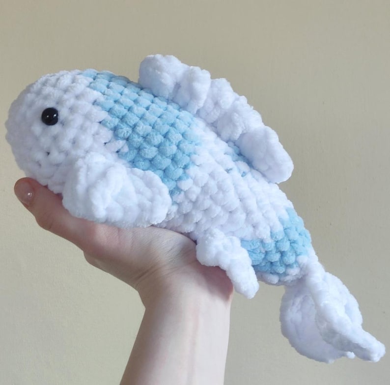 May include: A handmade, plush fish toy in white and light blue. The soft, crocheted fish has a rounded body with a blue pattern down its back and white fins. The toy has a black eye and is held in a hand.