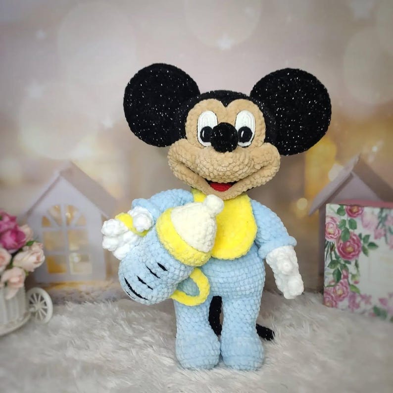 Puede incluir: Mu&ntilde;eco de Mickey Mouse hecho a mano con ganchillo, vistiendo un traje azul claro con un babero amarillo. El mu&ntilde;eco tiene orejas negras, cara color canela y sostiene un biber&oacute;n azul y amarillo. El mu&ntilde;eco est&aacute; sobre una superficie blanca y esponjosa.