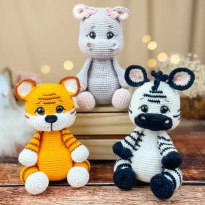 Pode incluir: Três brinquedos de animais de crochê feitos à mão: um tigre, um hipopótamo e uma zebra. O tigre é laranja com listras brancas e pretas. O hipopótamo é cinza com um laço rosa. A zebra é branca com listras pretas.