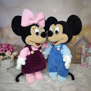 Puede incluir: Dos peluches de ratones, una pareja. La ratona tiene un lazo rosa, una camisa rosa y un vestido con peto morado oscuro. El rat&oacute;n lleva un peto azul. Ambos tienen orejas negras, caras color canela y guantes blancos.